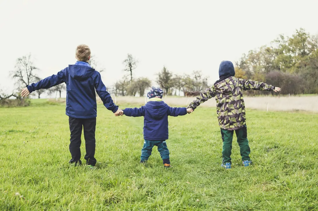 Trois enfants de dos se tiennent la main dans un pré.
