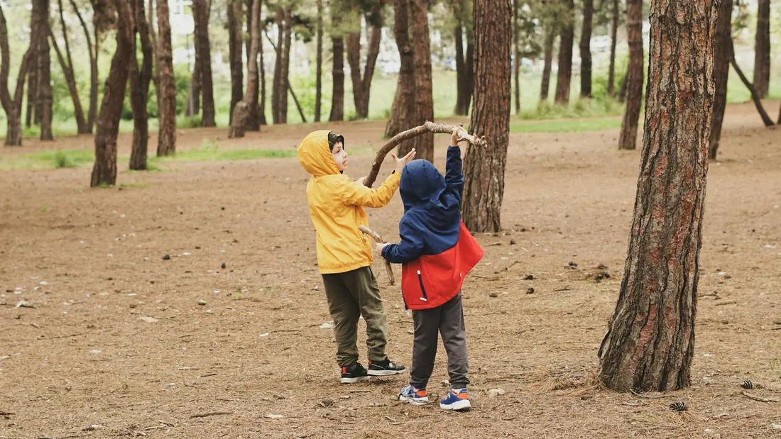 Deux enfants en imperméable jouent avec un bâton dans une pinède.